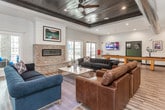 This single-floor community amenity space blends industrial and modern design elements to create a welcoming, multifunctional gathering environment. The main lounge area is a standout, featuring a dramatic tray ceiling finished in dark wood with a ceiling fan, a linear brick fireplace framed by shiplap, and an array of large windows that flood the space with natural light and provide views of the surrounding outdoor areas. Rich hardwood-style flooring runs throughout, anchoring plush navy blue sofas and leather seating arranged around a central coffee table for a comfortable, social atmosphere. A wall-mounted television adds to the entertainment options in this inviting lounge. The fully equipped kitchen is a highlight, showcasing dark cabinetry, a white subway tile backsplash, open wood shelving, pendant lighting, stainless steel appliances, and a large central island with bar seating — ideal for communal use and casual dining. A dedicated coffee and refreshment station adds everyday convenience. Adjacent to the lounge, a private dining area features a substantial wood table with upholstered seating, a wall-mounted display, and exposed industrial ductwork overhead that adds a contemporary, loft-inspired character. The space is thoughtfully designed to serve both social and functional needs.