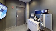 This contemporary lobby presents a refined, professional atmosphere defined by a cohesive palette of deep charcoal, cool grey, and brushed stainless steel. The entryway corridor leads visitors toward glass double doors at the front entrance, flanked by wall-mounted lighting that illuminates the polished tile flooring underfoot. Textured grey wall tiles and exposed concrete structural columns add architectural character and an industrial edge to the otherwise polished interior.

A standout feature is the stainless steel elevator with sleek panel doors, positioned prominently within the lobby and easily accessible from the main corridor. Nearby, a dedicated beverage station houses a Starbucks coffee and tea service, complete with a Tazo tea display, condiment setup, and a stainless steel coffee machine — providing a welcoming amenity for building occupants.

A wall-mounted flat-screen display serves as an informational hub, offering a functional and modern touch to the space. Dark accent walls contrast with white cabinetry along the refreshment counter, creating visual depth throughout. Safety and building systems, including a fire extinguisher and alarm pull station, are discreetly integrated into the design. Overall, the lobby conveys a polished, well-appointed environment suited to a professional commercial building.