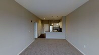 This single-floor unit offers a clean, open-concept layout that maximizes the sense of space and functionality. The living area features neutral-toned carpeting that adds warmth underfoot, while the smooth transition into the kitchen creates an effortlessly connected common space. The kitchen is a standout feature, showcasing a peninsula-style countertop with dark granite surfaces and a built-in sink, ideal for both meal preparation and casual dining. Stainless steel appliances, including a full-size refrigerator and range, bring a modern, polished aesthetic to the space. Above the peninsula, a pair of pendant lights provide focused illumination and a stylish design element against the neutral ceiling. White cabinetry lines the kitchen walls, offering ample storage. The flooring transitions to dark hardwood in the kitchen and entry areas, creating a clear visual distinction between zones. Closet doors with louvered panels are visible near the entryway, providing practical storage solutions. The neutral gray-toned walls throughout lend a contemporary, versatile backdrop suitable for a variety of furnishing styles. Recessed lighting and track lighting supplement the pendant fixtures, ensuring the space is well-lit throughout.
