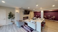 This single-floor communal space is thoughtfully designed with a contemporary aesthetic, blending comfort and functionality throughout its open layout. Light wood-look flooring flows seamlessly across the entire space, complementing a neutral color palette accented by rich burgundy-purple feature walls that add depth and visual interest.

The main lounge area serves as the focal point, anchored by a sleek floor-to-ceiling tiled fireplace wall with a linear electric fireplace and a large wall-mounted television above. Plush sectional seating, accent chairs, and a bold abstract area rug create an inviting gathering environment. Recessed lighting and wall sconces provide warm, layered illumination throughout.

Adjacent to the lounge, a bar-height counter with tufted red stools offers an additional social seating option. A coffee and refreshment station is conveniently positioned nearby, alongside a bold abstract artwork that reinforces the space's modern character.

The entryway features French-style glass doors that allow natural light to filter in, offering views of the outdoors. A small nook with French doors and casual floor cushions provides a quieter retreat within the space. The overall ambiance is polished yet approachable, making it well-suited for both relaxation and social gatherings.