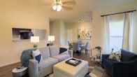 This single-story space showcases an open-concept layout that seamlessly connects the living and dining areas, creating a cohesive and functional environment. The living area is anchored by a comfortable neutral-toned sofa ensemble, complemented by an upholstered ottoman that doubles as a coffee table. A ceiling fan with an integrated light fixture overhead provides both airflow and ambient lighting, contributing to the room's welcoming atmosphere.

The flooring throughout features hardwood-style planks in a warm tone, adding visual continuity between spaces. A decorative area rug grounds the seating arrangement in the living area. Sheer white curtains frame a window, allowing soft natural light to filter into the space.

Transitioning into the dining area, a round table is surrounded by upholstered chairs in a complementary blue-grey palette. Track lighting above ensures the dining space is well-lit. Two framed abstract artworks adorn the wall, adding an artistic focal point. The warm beige walls and wainscoting detail in the dining area lend a refined, polished character to the overall interior design.