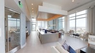 This stunning high-rise residence offers a seamlessly integrated open-concept living space defined by clean lines, contemporary finishes, and an abundance of natural light. Floor-to-ceiling windows wrap the interior, framing sweeping panoramic city views and flooding the space with daylight throughout the day.

At the heart of the home is a sleek, modern kitchen anchored by a large island with a polished countertop surface, providing ample workspace and an ideal setting for casual dining or entertaining. The kitchen's warm wood-accented ceiling detail, illuminated with subtle ambient lighting, adds a refined contrast to the otherwise cool, neutral palette.

The open layout flows effortlessly into a dedicated dining area, where a contemporary pendant light fixture adds a touch of elegance above the table. Beyond, the living area offers a relaxed yet sophisticated atmosphere, with plush seating and soft textiles that complement the light tile flooring underfoot.

The overall design aesthetic is modern and refined, with muted tones of white, grey, and warm wood creating a calm and polished ambiance. Thoughtful architectural ceiling detailing and recessed lighting enhance the sense of volume and depth throughout. This is a space that balances luxurious design with functional, comfortable living at an elevated level.