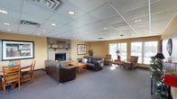 Located at 6495 Piping Rock Ln in Indianapolis, Indiana, this single-story space offers a well-appointed and inviting interior designed for comfort and community gathering. The main lounge area is a standout feature, anchored by a classic full-height brick fireplace with a wood mantel that serves as a warm focal point for the room. The space is furnished with dark leather sofas and upholstered armchairs arranged around a central coffee table, creating a relaxed and welcoming atmosphere. A row of large windows along one wall floods the room with natural light, offering pleasant outdoor views and enhancing the open, airy feel of the interior. The drop-tile ceiling features recessed lighting throughout, providing consistent, even illumination across the carpeted floor. Warm yellow-toned walls complement the traditional design elements, contributing to the overall cozy and approachable ambiance. The layout is thoughtfully organized to accommodate both intimate conversation areas and broader group gatherings, making it a functional and versatile space. The combination of classic architectural details, comfortable furnishings, and abundant natural light makes this a welcoming environment suited for a variety of uses.