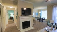 This single-floor residence offers a cohesive open-concept layout that connects the living and dining areas in a comfortable, flowing arrangement. The living space is anchored by a classic white-mantled fireplace, which serves as the room's focal point and doubles as a media wall with a large flat-screen television mounted above. Warm beige carpet runs throughout, complementing the neutral wall tones and white crown molding that frame the space with a traditional finish.

A gallery-style wall displaying framed artwork lines the hallway leading from the living area, adding a personal and curated touch. The dining area is positioned adjacent to the living room and is illuminated by a pendant light fixture hanging above the dining table, which is surrounded by dark wood chairs. Natural light filters through a window nearby, brightening the space and providing an airy feel.

Aqua and teal accents appear consistently throughout — from window treatments and artwork to decorative accessories — creating a unified color palette that adds vibrancy against the neutral backdrop. The hallway visible from the main living area hints at additional rooms beyond, suggesting a practical and well-organized floor plan throughout.