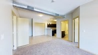 This single-floor space showcases an open-concept layout that seamlessly connects the living area, kitchen, and bathroom. The living area is finished with neutral-toned carpet, creating a warm and comfortable atmosphere, while the transition to the kitchen features light-colored tile flooring. An exposed metallic HVAC duct runs prominently across the ceiling, lending an industrial character to the space and adding visual interest overhead.

The kitchen is defined by dark-stained cabinetry that contrasts with the lighter walls and flooring. A peninsula-style counter provides additional workspace and subtle separation between the kitchen and living areas. Pendant lighting hangs above the kitchen, complementing the flush-mount ceiling fixtures that illuminate the open space.

A closet with sliding doors is accessible from the main living area, offering practical storage. The bathroom, visible from the central space, is finished with light-toned flooring and includes standard fixtures. An additional door leads to a separate room or closet area, adding versatility to the floor plan. The overall ambiance is clean, functional, and grounded in a contemporary industrial aesthetic.