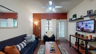 This single-floor unit is designed for functional, comfortable living, with each area thoughtfully arranged to maximize use of the available space. The living room serves as the heart of the home, featuring rich hardwood-style flooring, a bold terracotta accent wall, and a ceiling fan with integrated lighting. A window and a glass-paneled door allow natural light to filter in, while white drapes and plantation shutters provide privacy. Seating is anchored by a navy sofa and matching armchair arranged around a wooden coffee table.

The kitchen is well-equipped with warm wood cabinetry, dark countertops, white appliances including a range, microwave, and dishwasher, and a double sink. Track lighting illuminates the workspace, and a wooden dining table with black chairs offers a dedicated dining area nearby.

The bedroom continues the terracotta accent wall theme, complemented by carpeted flooring, a ceiling fan, and a window with blinds, creating a cozy and private retreat. A dedicated workspace with a desk is also present.

The bathroom features wood-toned cabinetry, a vanity with a mirror, and hardwood-style flooring. A dedicated laundry closet houses a full-size washer and dryer with overhead shelving for added storage.
