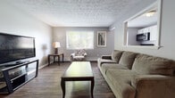 This single-floor space offers a thoughtfully arranged layout that balances comfort and practicality. The open-concept living and dining area is anchored by rich, dark hardwood-style flooring that flows throughout the main living spaces, complemented by neutral gray walls and a textured ceiling that adds subtle visual character. Large windows fitted with horizontal blinds allow natural light to fill the room, creating a bright and welcoming atmosphere.

The living area provides ample room for a full sofa arrangement, accent chair, and entertainment setup, with a dedicated dining nook accommodating a compact drop-leaf table and chairs. A framed piece of wall art adds a decorative touch to the dining corner.

The kitchen is efficiently designed with white cabinetry extending to the upper walls, offering generous storage. Black appliances — including a range, dishwasher, over-the-range microwave, and refrigerator — provide a clean contrast against the white cabinetry and light countertops. The dark hardwood-style flooring continues here, maintaining visual continuity.

The bedroom features plush carpeting underfoot, a window with outdoor greenery views, and a baseboard heating unit. The space comfortably accommodates a queen-sized bed and full dresser. The bathroom includes a tiled tub-shower combination, wood-look vinyl flooring, and clean white fixtures.