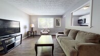 This single-floor space offers a thoughtfully arranged layout that balances comfort and practicality. The open-concept living and dining area is anchored by rich, dark hardwood-style flooring that flows throughout the main living spaces, complemented by neutral gray walls and a textured ceiling that adds subtle visual character. Large windows fitted with horizontal blinds allow natural light to fill the room, creating a bright and welcoming atmosphere.

The living area provides ample room for a full sofa arrangement, accent chair, and entertainment setup, with a dedicated dining nook accommodating a compact drop-leaf table and chairs. A framed piece of wall art adds a decorative touch to the dining corner.

The kitchen is efficiently designed with white cabinetry extending to the upper walls, offering generous storage. Black appliances — including a range, dishwasher, over-the-range microwave, and refrigerator — provide a clean contrast against the white cabinetry and light countertops. The dark hardwood-style flooring continues here, maintaining visual continuity.

The bedroom features plush carpeting underfoot, a window with outdoor greenery views, and a baseboard heating unit. The space comfortably accommodates a queen-sized bed and full dresser. The bathroom includes a tiled tub-shower combination, wood-look vinyl flooring, and clean white fixtures.