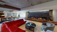 Located at 1120 N Division St in Spokane, Washington, this single-floor commercial space delivers a sophisticated industrial-modern aesthetic that makes a lasting impression from the moment of entry. The grand lobby sets the tone with a wide, carpet-runner corridor flanked by exposed concrete columns, warm wood-accented walls, and a stylish reception desk finished in natural wood and stone. Dramatic woven sphere pendant lights hang overhead, adding an artistic focal point against the high ceilings.

Moving deeper into the space, a sprawling lounge area commands attention with its bold red modular seating, a sleek round coffee table, and a long linear fireplace set against a floor-to-ceiling stacked slate surround — creating a warm, inviting atmosphere. Adjacent to the lounge, a formal meeting or dining area features a substantial hardwood table surrounded by contemporary chairs, anchored by a striking floral-patterned area rug and illuminated by industrial-style pendant lights. Large windows flood the area with natural light and offer views of the surrounding exterior.

Throughout the space, design details such as a living green wall, exposed concrete ceilings, reclaimed wood accent panels, and curated artwork contribute to a cohesive, upscale ambiance. Practical amenities including dual water fountains are also thoughtfully integrated.
