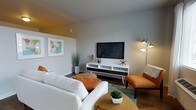 This single-floor residence showcases a thoughtfully designed interior that blends mid-century modern aesthetics with contemporary comfort. The living area serves as the heart of the space, anchored by rich hardwood-style flooring that adds warmth and depth throughout. Neutral gray walls provide a clean, versatile backdrop, complemented by striking pops of burnt orange in the furnishings — including plush throw pillows and a sleek accent chair with ottoman — that inject energy and personality into the room. A white and walnut media console with tapered legs serves as a focal point, housing a wall-mounted flat-screen television and decorative accents including coastal-inspired starfish and teal glassware. A pair of framed art prints adorns the wall, adding color and visual interest. The seating arrangement centers around a comfortable white sofa and a round wooden coffee table, creating an inviting and functional gathering space. Lush potted greenery and a modern arc floor lamp round out the ambiance, contributing to a bright and lively atmosphere. The overall design reflects a cohesive, curated style that balances retro-inspired character with modern livability.