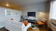 This single-floor residence showcases a thoughtfully designed interior that blends mid-century modern aesthetics with contemporary comfort. The living area serves as the heart of the space, anchored by rich hardwood-style flooring that adds warmth and depth throughout. Neutral gray walls provide a clean, versatile backdrop, complemented by striking pops of burnt orange in the furnishings — including plush throw pillows and a sleek accent chair with ottoman — that inject energy and personality into the room. A white and walnut media console with tapered legs serves as a focal point, housing a wall-mounted flat-screen television and decorative accents including coastal-inspired starfish and teal glassware. A pair of framed art prints adorns the wall, adding color and visual interest. The seating arrangement centers around a comfortable white sofa and a round wooden coffee table, creating an inviting and functional gathering space. Lush potted greenery and a modern arc floor lamp round out the ambiance, contributing to a bright and lively atmosphere. The overall design reflects a cohesive, curated style that balances retro-inspired character with modern livability.