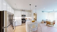This thoughtfully designed single-floor residence offers a clean, contemporary aesthetic anchored by light hardwood flooring and a bright, neutral color palette throughout. The open-concept kitchen is a standout feature, outfitted with full-height white shaker cabinetry, quartz countertops, stainless steel appliances — including a refrigerator, range, microwave, and dishwasher — and a central island with bar seating. Dual pendant lights with clear glass shades hang above the island, complemented by track lighting overhead. The adjacent living area is airy and well-lit, featuring exposed metallic ductwork that adds an industrial accent to the otherwise minimalist design. Large windows and a glass balcony door flood the space with natural light and provide outdoor access with urban views. The primary bedroom is generously sized with a walk-in closet and connects to a full bathroom featuring a double-sink vanity, white cabinetry, and framed mirrors. A second bedroom offers a comfortable, quiet retreat, and a second full bathroom includes a combined tub and shower with a single-sink vanity. The overall ambiance is clean, modern, and move-in ready.