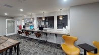 This single-floor business center and lounge is thoughtfully designed to support both productivity and relaxed collaboration. The space is finished with a neutral gray color palette, crisp white trim, and a patterned commercial carpet in charcoal tones that grounds the room with a contemporary feel.

Along one wall, a continuous wall-mounted desk provides a dedicated computer workstation area, equipped with multiple desktop computers, stylish wood-and-chrome swivel chairs, and a series of decorative gold pendant lights that hang overhead, adding warmth to the functional setup. Large windows above the workstation counter allow natural light to filter in while offering views of the surrounding exterior.

A central gathering area features a rustic wood communal table paired with bold yellow accent chairs — a design choice that injects energy and personality into the space. A wall-mounted flat-screen television is positioned against a textured accent wall, creating an inviting lounge corner ideal for informal meetings or casual use.

Floor-to-ceiling glass-panel doors open toward an outdoor deck, further enhancing the sense of openness and bringing in additional natural light. Recessed ceiling lighting provides even illumination throughout. A private glass-door room along one side offers a more enclosed area for focused work or quiet use, completing this well-appointed and versatile community workspace.