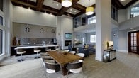This single-floor space makes a striking impression with its dramatic double-height ceilings accented by rich dark wood coffered beams and elegant pendant lighting, including a bold yellow drum shade that adds a pop of color. The open-plan layout seamlessly connects multiple functional zones, creating a cohesive yet versatile environment.

A central dining area anchors the space, featuring a substantial warm-toned wooden table surrounded by modern high-back chairs in contrasting dark and white finishes. Adjacent to it, a wet bar with a mosaic tile backsplash, dark cabinetry, and barstool seating serves as a social hub, complete with a coffee station.

The lounge area offers plush gray upholstered sofas and accent chairs arranged around a sleek built-in fireplace, with a wall-mounted flat-screen TV above it for entertainment. Natural light filters in through windows at the far end, illuminating the neutral-toned flooring that ties the space together.

An upper mezzanine-level window detail adds architectural depth, while recessed lighting throughout ensures a well-lit, welcoming ambiance at all hours.