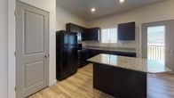 This single-floor residence offers a thoughtfully designed layout with a cohesive, modern aesthetic throughout. The kitchen serves as a functional centerpiece, featuring rich dark cabinetry paired with light granite countertops that provide striking visual contrast. Black appliances, including a refrigerator, electric range, microwave, and dishwasher, complement the cabinetry, while light hardwood-style flooring adds warmth underfoot. A peninsula counter extends the workspace and enhances the kitchen's utility. Natural light filters in through a window fitted with blinds, brightening the space.

The bedroom is finished with wall-to-wall carpet in a neutral tone, lending a comfortable and quiet atmosphere. A ceiling fan with a light fixture provides both climate control and illumination. The clean white walls and simple trim throughout maintain a consistent, understated palette.

The bathroom is finished with stone-look tile flooring and features a full tub-and-shower combination with built-in shelving, a dark-cabineted vanity with a granite countertop, and recessed lighting. A utility room houses a water heater, HVAC system, and stacked washer and dryer, consolidating essential mechanical systems and in-unit laundry into one organized space. Overall, the unit presents a clean, functional living environment with modern finishes.