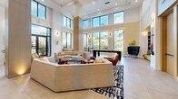 Located at 8300 Clearly Blvd in Plantation, Florida, this single-floor community living space is a thoughtfully designed gathering area that blends contemporary style with functional amenities. The space is anchored by dramatically high ceilings that amplify the sense of openness, while floor-to-ceiling windows along multiple walls flood the interior with natural light and offer views of the surrounding palm-lined outdoor areas. Large-format neutral tile flooring runs throughout, providing a clean and cohesive foundation for the varied zones within.

The central lounge area features generously sized, curved neutral sofas and warm cognac leather accent chairs arranged around richly toned wooden coffee tables and stools, all set atop a bold navy and white abstract area rug. A wall-mounted flat-screen television is framed by a warm wood panel backdrop and flanked by pendant lighting, creating an inviting media and social hub. Adjacent to this is a dedicated game zone housing a substantial billiards table, complemented by built-in shelving and seating.

A sleek coffee bar station with gray cabinetry, a white quartz countertop, and stylish pendant lighting offers a convenient self-serve refreshment area. The overall ambiance is bright, welcoming, and sophisticated—ideal for socializing, relaxing, or recreation.