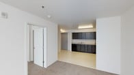 This single-floor space presents a clean, open layout designed for functionality and ease of use. The interior is defined by crisp white walls and smooth white ceilings, creating a bright and neutral atmosphere throughout. The space transitions between two distinct flooring types — a warm, beige carpet covers the main living or gathering area, while a light-toned vinyl or linoleum floor extends into the kitchenette zone, clearly delineating the two areas.

The kitchenette is equipped with dark-toned cabinetry lining the wall, offering generous upper and lower storage. A built-in sink with a countertop workspace sits beneath fluorescent strip lighting, providing ample illumination for the work area. The dark cabinetry creates a bold contrast against the light walls and floors, adding a modern touch to an otherwise minimal design.

A set of double doors with frosted glass panels leads to an adjacent room or closet space, adding a refined architectural detail. A secondary solid door is also visible, providing access to additional areas. Recessed sprinkler heads and a smoke detector are integrated into the ceiling, reflecting a practical and safety-conscious design. Overall, the space offers a versatile, well-organized layout suitable for a range of uses.