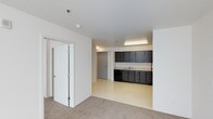 This single-floor space presents a clean, open layout designed for functionality and ease of use. The interior is defined by crisp white walls and smooth white ceilings, creating a bright and neutral atmosphere throughout. The space transitions between two distinct flooring types — a warm, beige carpet covers the main living or gathering area, while a light-toned vinyl or linoleum floor extends into the kitchenette zone, clearly delineating the two areas.

The kitchenette is equipped with dark-toned cabinetry lining the wall, offering generous upper and lower storage. A built-in sink with a countertop workspace sits beneath fluorescent strip lighting, providing ample illumination for the work area. The dark cabinetry creates a bold contrast against the light walls and floors, adding a modern touch to an otherwise minimal design.

A set of double doors with frosted glass panels leads to an adjacent room or closet space, adding a refined architectural detail. A secondary solid door is also visible, providing access to additional areas. Recessed sprinkler heads and a smoke detector are integrated into the ceiling, reflecting a practical and safety-conscious design. Overall, the space offers a versatile, well-organized layout suitable for a range of uses.