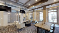 This single-story clubhouse is a well-appointed community space designed to foster connection and relaxation. The interior is defined by soaring coffered ceilings with rich, dark wood detailing that lend a refined, architectural character to the room. Decorative columns frame the open layout, adding a classic, stately feel while preserving a sense of openness throughout.

The main gathering area features a variety of thoughtfully arranged seating options, including plush lounge chairs and low-profile coffee tables arranged atop a patterned area rug, creating an inviting atmosphere for casual conversation or quiet lounging. A wall-mounted flat-screen television is centrally positioned, making the space equally suited for communal entertainment.

Large windows along the perimeter allow generous natural light to fill the interior, offering views of the greenery outside and enhancing the warm, welcoming ambiance. A communal high-top wooden table adds a rustic contrast to the more formal architectural elements, providing an additional space for dining or socializing.

Toward the rear of the space, a service counter or bar area is visible, suggesting amenities for residents and guests. Wall sconces complement the recessed lighting, contributing to a layered and inviting illumination scheme. Overall, this clubhouse strikes a balance between elegance and approachability, making it a versatile and attractive community hub.