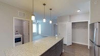 Located at 4001 Reedybrook Xing in Apex, NC, this single-floor residence offers a thoughtfully designed open-concept layout that blends style and functionality. The kitchen serves as a central highlight, anchored by a generous granite-topped island with a light, veined finish and dark gray cabinetry accented by brushed nickel hardware. Three pendant lights hang above the island, providing both task lighting and a refined aesthetic touch. The kitchen flows seamlessly into the living area, which is brightened by large windows that invite natural light and include a ceiling fan for year-round comfort. Durable wood-look flooring extends throughout the main living spaces, complementing the neutral gray and white color palette that gives the home a clean, contemporary feel. A walk-in closet with built-in shelving offers ample storage, while additional doors throughout suggest well-appointed utility and bedroom spaces. The overall ambiance is modern and move-in ready, with crisp white trim, smooth ceilings, and a cohesive design that makes this single-level home both practical and visually appealing.
