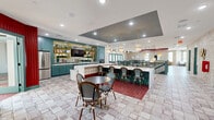 Located at 8052 Thayer Way in West Chester, Ohio, this expansive single-floor amenity space offers a thoughtfully designed environment for socializing, recreation, and everyday convenience. The layout spans a generous footprint, connecting a series of distinct functional areas through a cohesive and boldly styled interior aesthetic.

The open kitchen and living area serves as the centerpiece of the space, featuring striking teal cabinetry with brass hardware, sweeping white quartz countertops, and decorative gold open shelving. Patterned tile flooring, a coffered dark ceiling inset with recessed lighting, and pendant fixtures add layers of visual interest. A large island with bar seating makes this an ideal gathering hub.

Adjacent living areas showcase exposed light brick walls, herringbone hardwood-style flooring, and a dramatic coffered tin ceiling with dark framing. Comfortable lounge seating surrounds a shuffleboard table, while warm pendant lighting and bold red ceiling accents create an inviting atmosphere. A separate seating nook with floor-to-ceiling teal-framed glass doors offers a more intimate retreat.

A dedicated recreation room houses a golf simulator with turf putting surface, exposed brick walls, and a coffered ceiling with grid detailing, all bathed in natural light from multiple windows. The hallway connects the zones with clean lines, teal railings, and a bold red accent wall. A laundry facility rounds out the practical offerings of this well-appointed community space.