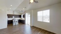 This single-story space showcases a thoughtfully designed open-concept layout that seamlessly connects the kitchen and living areas. The kitchen is outfitted with dark espresso cabinetry that provides a striking contrast against the crisp white walls, and includes stainless steel appliances such as a microwave and oven. A peninsula with bar seating accommodates metal stools, offering a casual dining option. Rich wood-look flooring extends throughout the space, adding warmth and continuity to the design. Vaulted ceilings enhance the sense of openness, and a ceiling fan with integrated lighting serves both the functional and aesthetic needs of the room. Natural light flows generously into the space through large windows fitted with white horizontal blinds, as well as a sliding glass door that leads to an outdoor area, creating a bright and airy atmosphere. The neutral white walls provide a versatile backdrop, keeping the overall ambiance clean, modern, and welcoming. The combination of contemporary finishes and practical design elements makes this a highly functional and visually appealing space.