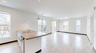 Located at 1136 E North Ave in Milwaukee, WI, this modern 2-bedroom, 2-bathroom unit offers a thoughtfully designed open-concept floor plan finished with light hardwood-style flooring throughout the main living areas. The kitchen serves as a central hub, featuring light wood cabinetry, stainless steel appliances including a refrigerator, gas range, and microwave, a large island with an integrated sink and countertop seating, and pendant lighting overhead. The kitchen flows seamlessly into a bright, spacious living room bathed in natural light from oversized windows and sliding glass doors that open onto a private balcony with views of the surrounding neighborhood. The primary bedroom is generously sized, offering a sliding glass door to the balcony, additional windows for abundant natural light, and direct access to its own full bathroom. The second bedroom features plush carpeting, dual windows with urban views, and a sliding closet with a clothes rail. Both bathrooms are clean and contemporary — one with a tub/shower combo and the other with a walk-in shower and light wood vanity storage. A dedicated in-unit laundry closet with a full-size washer adds everyday convenience. The overall ambiance is bright, clean, and modern throughout.