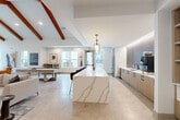 Located at 700 Avett Ln in Charlotte, NC, this single-floor community space offers a thoughtfully designed open-concept layout that seamlessly blends a fully equipped kitchen with an expansive living and recreation area. The space is immediately distinguished by its dramatic vaulted ceilings adorned with rich wood beams, creating an airy, loft-like atmosphere throughout.

The living area serves as the focal point, anchored by a floor-to-ceiling brick fireplace flanked by large windows that invite abundant natural light. Comfortable seating arrangements featuring cognac leather chairs and neutral-toned sofas are thoughtfully positioned across herringbone-patterned wood flooring, while a billiards table adds a recreational element to the space.

The kitchen area is equally impressive, centered around a striking waterfall-edge marble island with bar seating and a geometric pendant light overhead. The galley-style kitchen runs along one wall, featuring light wood cabinetry, white quartz countertops, a bold hexagonal dark tile backsplash with LED accent lighting, stainless steel appliances, and a dedicated coffee station. The cohesive design palette of warm neutrals, natural wood tones, and modern finishes gives the entire space a welcoming yet sophisticated character ideal for gathering and socializing.