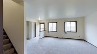This 3-floor property offers a well-designed layout with clean, neutral interiors that create a bright and welcoming atmosphere. The main living area is generously sized, featuring soft wall-to-wall carpeting in a light grey tone that adds warmth and comfort underfoot. Crisp white walls and a smooth ceiling contribute to the open, airy feel of the space.

Multiple windows with dark-trimmed frames allow abundant natural light to fill the room, offering views of the surrounding exterior. Sheer curtains provide a soft finishing touch while maintaining brightness. A half-wall or breakfast bar element subtly defines the transition between the living area and an adjacent space, adding architectural interest without closing off the floor plan.

A closet with open shelving is visible toward the back of the room, providing practical storage solutions. The flooring transitions hint at distinct functional zones within the layout. Baseboard heating units are present along the walls, ensuring comfort across seasons.

Spanning three floors, the property provides ample room for varied living arrangements, with each level likely offering its own distinct spaces and functionality suited to everyday living.