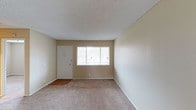 This single-story space presents a straightforward and functional layout, highlighted by a well-maintained interior with neutral, beige-toned walls that create a warm and versatile backdrop. The main living area is finished with wall-to-wall carpet in a light tan hue, providing a soft underfoot feel while contributing to the room's understated, clean aesthetic. Near the entryway, a transition to hardwood-style flooring adds a subtle contrast and defines the entry point of the space.

A white entry door with a standard lock provides access to the unit, positioned adjacent to a centrally placed window fitted with horizontal blinds. This window allows a notable amount of natural light to filter into the room, brightening the interior and lending an open, airy quality to the space. The textured popcorn ceiling adds a classic residential character throughout.

The walls are clean and unadorned, offering ample flexibility for personalization and furniture arrangement. White baseboards frame the perimeter of the room neatly, contributing to a tidy, cohesive finish. Overall, the space is move-in ready, presenting a simple, functional environment with a neutral palette suited to a wide range of uses.