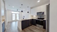 This contemporary single-floor unit showcases a thoughtfully designed open-concept layout that seamlessly connects the kitchen, dining, and living areas. The kitchen is a standout feature, boasting two-tone cabinetry with dark lower cabinets and crisp white upper cabinets, granite countertops, a subway tile backsplash, and a full suite of stainless steel appliances including a range, microwave, dishwasher, and refrigerator. A peninsula with an undermount sink and matte black faucet doubles as a breakfast bar, while two sleek pendant lights add a modern, industrial touch overhead. Wood-look plank flooring runs throughout the main living areas, complementing the clean, neutral color palette. The living area is bathed in natural light from French doors that open to a private balcony, with a ceiling fan adding to the airy ambiance. The bedrooms feature plush carpeting, ceiling fans, and generous windows with blinds that allow for both natural light and privacy. The bathroom is elegantly finished with a navy blue vanity, granite countertop, an illuminated mirror, matte black hardware, and a full soaking tub with tile surround. Throughout the unit, clean lines, contemporary fixtures, and a cohesive design aesthetic create a polished and inviting living environment.