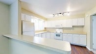 This single-floor home offers a well-designed layout centered around a generously sized kitchen that serves as the heart of the space. The kitchen is outfitted with an abundance of white raised-panel cabinetry that extends to the upper walls, providing ample storage throughout. A large, light-colored countertop breakfast bar anchors the space, offering both a functional workspace and a casual dining area. The kitchen is equipped with white appliances, including a refrigerator, electric range, built-in microwave, and dishwasher, creating a cohesive and clean aesthetic. A double-basin sink is positioned beneath a multi-pane window that invites natural light deep into the room. Track lighting along the ceiling supplements the natural light, ensuring the space remains bright and well-illuminated at all times. The warm yellow-toned walls complement the white cabinetry and light countertops, lending the kitchen a welcoming and airy ambiance. Dark hardwood flooring grounds the space with a rich contrast against the lighter elements above. The overall design emphasizes functionality and openness, making this kitchen a practical and inviting environment for everyday living and entertaining.