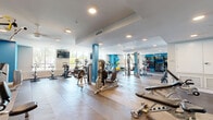 Located at 3675 T St in Sacramento's 95816 zip code, this well-appointed fitness center spans a generous single floor, offering an extensive and thoughtfully designed workout environment. The centerpiece is a large, open exercise room distinguished by its herringbone-patterned hardwood flooring, recessed lighting, and ceiling fans that keep the space comfortable during intense workouts. Bold teal accent walls and columns add a vibrant pop of color against the crisp white walls and ceilings, creating an energetic yet modern aesthetic. Large windows along multiple walls flood the space with abundant natural light and provide views of the surrounding Sacramento neighborhood. The exercise room is comprehensively equipped with a wide variety of cardio machines — including treadmills, ellipticals, stationary bikes, and rowing equipment — alongside a robust selection of strength training machines, cable systems, free weights, and functional fitness accessories such as medicine balls, stability balls, and resistance tools. A dedicated wall-mounted equipment rack keeps accessories organized. Wall-mounted TVs enhance the cardio experience. The facility also includes two ADA-compliant bathrooms, each finished with clean white subway tile, grab bars, and warm wood-accented vanities, ensuring accessibility and comfort for all users.