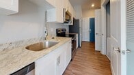 This single-floor unit offers a well-designed layout with consistent gray-toned hardwood flooring throughout, lending a cohesive and modern aesthetic to the entire space. The interior is finished with neutral gray walls and crisp white trim, creating a clean, contemporary atmosphere.

The main living area is generously proportioned and filled with natural light from a large sliding glass door that opens onto a private balcony, providing pleasant outdoor views of the surrounding neighborhood. Recessed lighting complements the natural light in other areas of the unit.

The kitchen is thoughtfully outfitted with white shaker-style cabinetry, light granite countertops featuring subtle veining, a stainless steel single-basin sink with a modern pull-down faucet, and a full suite of stainless steel appliances including a refrigerator, electric range, over-the-range microwave, and dishwasher.

The bedroom includes a walk-in closet with louvered doors, offering ample storage. A louvered closet door is also present near the entry area.

The bathroom features a soaking tub with subway tile surround, a white shaker-style vanity with solid white countertop, an undermount sink, and a large frameless mirror, all finished in a clean, minimalist style.