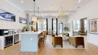 Located at 3450 S Blairstone Rd in Tallahassee, FL, this single-floor clubhouse-style space offers a thoughtfully designed interior that blends contemporary elegance with warm, inviting details. Light-toned hardwood flooring flows throughout, unifying each area seamlessly.

The entrance welcomes visitors with a round statement table, curated art, and a glimpse into the expansive open-plan interior beyond. The central living area features soaring ceilings with exposed beams, a bold brass chandelier, and an arrangement of plush velvet and leather seating around a marble-top coffee table, all set against sage-green paneled walls. Large floor-to-ceiling windows dressed in vibrant botanical-print drapes flood the space with natural light and provide outdoor views.

The adjacent lounge and dining zone centers around a light blue kitchen island with bar seating, pendant lighting, and a beverage station — ideal for social gatherings. A dedicated workspace nook includes a built-in desk along a shiplap-accented wall, outfitted with computers and a printer beneath tasteful framed artwork and warm sconce lighting.

A hallway connects to additional rooms, including a closet and transitional spaces, all finished with consistent, gallery-style wall art. The overall ambiance is sophisticated yet approachable, with a cohesive palette of whites, sage greens, and warm earth tones throughout.