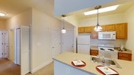 This single-floor residence is thoughtfully designed to maximize functionality and comfort. The kitchen serves as a central highlight, featuring warm honey-toned wooden cabinetry that provides ample storage alongside white appliances, including a full-size refrigerator and range. A stainless steel sink is seamlessly integrated into the countertop, adding a practical touch to the space. The kitchen opens to a peninsula-style breakfast bar with a light-toned countertop, ideal for casual dining or entertaining. Two elegant chain-hung pendant lights hang above the bar, casting warm, focused light over the surface. A skylight above the kitchen area floods the space with natural light, creating a bright and airy atmosphere. The open layout connects the kitchen to the adjacent living and entryway areas, which feature neutral carpet flooring and crisp white walls that enhance the sense of openness. Smooth, clean-lined white interior doors contribute to the cohesive aesthetic throughout. The overall ambiance is warm, functional, and inviting, with a design that balances everyday practicality with comfortable residential living.