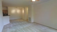 This single-floor space presents an open, interconnected layout finished in neutral, warm beige tones throughout. The main living area is carpeted with light-colored, plush carpeting that extends seamlessly across the room, contributing to a cohesive and understated aesthetic. The space is illuminated by two distinct lighting fixtures: a ceiling fan with an integrated light kit positioned centrally, and a contemporary track lighting rail mounted in the adjoining area, providing flexible, directional illumination. A floor-level HVAC vent is integrated into the wall, indicating a forced-air climate control system. The walls are smooth and painted in a consistent off-white or light beige palette, lending a clean, neutral backdrop throughout. A paneled interior door is visible toward the rear, suggesting additional rooms or utility spaces beyond the main living area. The layout opens broadly between the two visible zones, creating a generous sense of flow and usable space. The overall ambiance is clean, bright, and move-in ready, with a practical, straightforward design well-suited for a variety of uses.