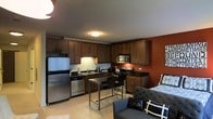 This single-floor space offers a thoughtfully designed open-concept layout that seamlessly integrates the kitchen and living area into one cohesive environment. The kitchen is a standout feature, outfitted with stainless steel appliances including a full-size refrigerator, gas range, built-in microwave, and dishwasher. Rich, dark wood cabinetry lines the walls from floor to ceiling, providing generous storage, while dark granite countertops add a polished, upscale feel. A bold terra cotta accent wall adds warmth and visual depth to the space, complementing the dark tones of the cabinetry. A compact dining area with a sleek metal table and bar-height stools sits at the heart of the room, making efficient use of the available space. The living area flows naturally from the kitchen, furnished with contemporary upholstered seating featuring decorative throw pillows, creating a comfortable and inviting atmosphere. Light tile flooring runs throughout, brightening the interior and providing a clean contrast to the darker elements. Flush-mount ceiling lighting ensures the space is well-lit throughout. Overall, the unit strikes a balance between functionality and style, making it a practical yet attractive living environment.