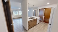 Located at 175 Pump Dr, Unit 103 in Burlington, Washington, this single-floor residence features a clean, functional layout with modern finishes throughout. The open-concept kitchen serves as the heart of the unit, equipped with warm wood cabinetry, a double-basin stainless steel sink set into a peninsula with light countertops, pendant lighting, a white refrigerator, electric range, built-in microwave, and a dishwasher. A conveniently located in-unit laundry closet is accessible directly from the kitchen area. The kitchen flows seamlessly into a spacious living area, which is bathed in natural light from large windows that frame views of the surrounding landscaped grounds and neighboring building exteriors. Light wood-tone luxury vinyl plank flooring extends throughout the unit, contributing to a cohesive and easy-to-maintain aesthetic. The bedroom offers a quiet, well-lit retreat with a window overlooking the greenery outside, while the bathroom features a wide vanity with warm wood cabinetry, a large mirror with overhead lighting, and a combined tub and shower. The neutral color palette of soft whites and warm wood tones creates a calm, inviting atmosphere throughout the entire space.