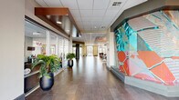 Located at 1141 W Stadium Dr in Fayetteville, Arkansas, this dynamic single-floor community clubhouse spans nearly 3,000 square feet of thoughtfully designed social and recreational space. The interior makes an immediate impression with a wide, light-filled entrance corridor adorned with large-scale contemporary artwork, potted tropical plants, and large glass storefronts that allow natural light to pour in.

The open floor plan transitions seamlessly between distinct activity zones. A central lounge area features bold orange drop ceilings, cylindrical steel columns, and an entertainment wall with a flat-screen TV framed in warm wood and brick, surrounded by stylish egg chairs, modular sofas, and colorful accent rugs. Adjacent to this is a game room equipped with a full-size billiard table, a dramatic pendant chandelier, and vibrant red accent walls.

A communal kitchen and bar area offers a large curved countertop with red leather barstools, pendant bottle-style lighting, a mosaic tile backsplash, stainless steel appliances, and a distinctive chartreuse ceiling. A high-top bar counter with industrial-style stools provides additional casual seating throughout the main floor.

Glass-walled fitness facilities are visible from the interior, with direct outdoor access through full-height glass doors. The overall design blends bold colors, mixed materials, and contemporary furnishings to create an energetic yet functional communal environment.