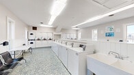 Located at 181 Derrer Rd in Columbus, Ohio, this community laundry facility offers a clean, bright, and efficiently organized space designed for convenience and comfort. The single-floor layout is thoughtfully arranged to maximize functionality, with a generous row of top-load washing machines and a full bank of dryers lining the walls, providing ample capacity for residents. A deep utility sink offers added practicality for hand-washing or pre-treating garments. Folding tables are positioned throughout the space, giving users plenty of room to sort and fold laundry with ease. The interior features a crisp white color palette accented by white beadboard wainscoting that runs along the lower walls, contributing to a clean and polished aesthetic. The speckled mosaic tile flooring in soft blue and gray tones adds a touch of character underfoot. Bright fluorescent lighting ensures excellent visibility throughout. Natural light filters in through multiple windows, creating an open and welcoming atmosphere. A dedicated waiting area with rows of seating allows residents to relax comfortably while their laundry cycles complete. The space is well-maintained and thoughtfully designed to serve as a reliable, shared amenity for the surrounding residential community.
