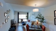 This single-floor space is thoughtfully designed with an open-plan layout that integrates the living and dining areas into one cohesive, flowing environment. The living area features light-toned walls that reflect natural light streaming in through a large window dressed with dark curtains, creating a bright and welcoming atmosphere. Wood-look flooring runs throughout, adding warmth and continuity across the space.

The living area is furnished with a comfortable white sofa accented by colorful throw pillows, accompanied by an eclectic patterned armchair that adds a touch of personality. A sleek media console anchors one wall, providing functional storage and display space. Lush indoor plants are strategically placed to introduce natural elements and vibrancy.

The dining area is defined by a modern glass-top table surrounded by upholstered chairs in rich orange tones, grounded beneath a stylish drum pendant light that serves as a focal point overhead. Place settings and decorative centerpieces on the dining table suggest a space designed for both everyday use and entertaining. Overall, the space presents a balanced blend of modern design and comfortable, livable style.