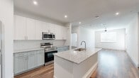 This thoughtfully designed single-floor residence offers a clean, contemporary aesthetic defined by crisp white walls, warm hardwood-style flooring, and recessed lighting throughout. The open-concept kitchen serves as the heart of the home, showcasing a large center island with a granite countertop and integrated sink, sage-toned shaker-style cabinetry, a white subway tile backsplash, and a full suite of stainless steel appliances including a range, over-the-range microwave, dishwasher, and French door refrigerator. An in-unit washer and dryer are conveniently tucked within the kitchen area. The kitchen flows seamlessly into a generous living area, illuminated by natural light from a large window and finished with a ceiling fan overhead. The bedroom offers ample space, a ceiling fan, and a well-sized window that brings in natural light. The primary bathroom features a granite-topped vanity with sage cabinetry, a tub-shower combination with white tile surround, and direct access to a walk-in closet equipped with multiple shelving rails. Additional rooms provide flexible space with matching flooring and bright, neutral finishes. The overall ambiance is modern, clean, and move-in ready.