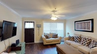 This single-story space presents a warm and inviting living area designed with comfort and functionality in mind. The living room serves as the focal point of the space, featuring rich, dark hardwood flooring that anchors the room and adds a sense of warmth throughout. Soft yellow walls create a bright, cheerful atmosphere, complemented by crisp white crown molding that adds a clean, polished finish to the space.

A large front window fitted with white horizontal blinds allows an abundance of natural light to fill the room, enhancing its open and airy feel. The dark front door, framed with classic white trim and adorned with a sheer curtain panel, provides a strong contrast to the light-toned walls and adds to the overall character of the space.

A ceiling fan with an integrated light fixture is centrally positioned overhead, offering both practical air circulation and ambient lighting. The layout comfortably accommodates multiple seating pieces, suggesting a well-proportioned living area suited for everyday use. A small accent piece near the entry wall adds a touch of functional décor, rounding out this welcoming and neatly appointed space.