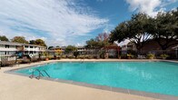 This amenity space features two distinct areas designed to support an active lifestyle. The outdoor pool area is generously sized, showcasing clear turquoise water surrounded by a spacious tiled deck. A wrought-iron perimeter fence encloses the area, complemented by mature shade trees, potted plants, and a pergola structure that adds architectural character. Seating and umbrella-shaded tables provide comfortable lounging options beneath open skies.

The fitness center is bright and modern, featuring light-toned carpet tile flooring, recessed lighting, and a ceiling fan that promotes air circulation throughout the space. Floor-to-ceiling mirrors line the walls, amplifying the sense of openness and allowing users to monitor their form. Large windows and sliding glass doors invite abundant natural light and offer direct views of the pool area, creating a seamless indoor-outdoor connection.

Equipment is well-rounded and includes multiple treadmills, elliptical machines, and a stationary bike for cardio training. Strength training is supported by a multi-station cable machine, adjustable benches, a full dumbbell rack, and a medicine ball storage tower. Open shelving provides organized storage for accessories such as resistance tools and rolled towels, while a wall-mounted television adds entertainment during workouts.