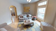 This charming single-floor residence exudes a warm and inviting atmosphere, thoughtfully designed for comfortable everyday living. The living room serves as the heart of the space, showcasing beautiful light hardwood floors that run throughout and large windows that flood the room with abundant natural light. A neutral-toned sofa anchors the seating area, complemented by an upholstered ottoman coffee table and a patterned area rug featuring yellow floral accents that add a cheerful pop of color. A decorative world map canvas adorns the wall, lending a well-traveled, personalized character to the room. A distinctive arched doorway connects the living area to the adjoining hallway, adding an elegant architectural detail that enhances the overall charm of the home. The soft, neutral color palette throughout creates a calm and airy ambiance, while thoughtful décor choices — including woven baskets and accent pillows in teal and coral — bring warmth and personality to the space. The layout flows naturally between rooms, making the home feel open and connected. Overall, this residence offers a comfortable and stylish living environment with classic architectural touches and a welcoming, light-filled atmosphere.