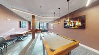 This dedicated game room offers a thoughtfully designed recreational space finished with a cohesive modern aesthetic. The room is dressed in warm mauve and terracotta-toned walls, complemented by herringbone-patterned carpet tiles that add visual texture underfoot. Linear LED lighting runs across the ceiling in dynamic angular arrangements, providing bright, even illumination throughout the space, while pendant lights with glass shades hang above the central pool table, adding a stylish focal point.

The room is well-equipped for entertainment, housing a full-size pool table with a wall-mounted cue rack nearby, a long shuffleboard table positioned along one wall, and a foosball table occupying a dedicated corner. Framed artwork and a large display screen mounted on the wall contribute to the room's polished, curated atmosphere. A cluster of dark leather chairs offers comfortable seating for spectators.

Large windows along one wall invite natural light and provide outdoor views, enhancing the openness of the space. Access is provided through sleek glass double doors, maintaining visual connectivity to the adjacent corridor. Overall, the room balances a lively, social atmosphere with clean, contemporary design details.