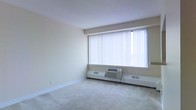 This single-floor space offers a clean, functional layout with a neutral aesthetic throughout. The main living area is finished with wall-to-wall carpet in a light beige tone, contributing to a warm and quiet atmosphere underfoot. A prominent, large window dressed with vertical blinds serves as the focal point of the room, allowing an abundance of natural light to fill the interior while offering adjustable privacy and light control. Beneath the window, a wall-mounted PTAC (packaged terminal air conditioner) unit provides convenient, self-contained climate control, supplemented by baseboard heating units that run along the perimeter of the room. The walls are finished in a soft, neutral off-white tone, and the ceiling features a smooth, consistent texture that keeps the visual space open and uncluttered. White baseboards frame the flooring cleanly throughout. The overall design is straightforward and practical, presenting a blank canvas well-suited for a variety of uses. The abundance of natural light from the large window enhances the sense of openness in the room, making the space feel airy and inviting despite its understated simplicity.
