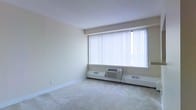 This single-floor space offers a clean, functional layout with a neutral aesthetic throughout. The main living area is finished with wall-to-wall carpet in a light beige tone, contributing to a warm and quiet atmosphere underfoot. A prominent, large window dressed with vertical blinds serves as the focal point of the room, allowing an abundance of natural light to fill the interior while offering adjustable privacy and light control. Beneath the window, a wall-mounted PTAC (packaged terminal air conditioner) unit provides convenient, self-contained climate control, supplemented by baseboard heating units that run along the perimeter of the room. The walls are finished in a soft, neutral off-white tone, and the ceiling features a smooth, consistent texture that keeps the visual space open and uncluttered. White baseboards frame the flooring cleanly throughout. The overall design is straightforward and practical, presenting a blank canvas well-suited for a variety of uses. The abundance of natural light from the large window enhances the sense of openness in the room, making the space feel airy and inviting despite its understated simplicity.