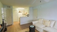 This single-floor space features an open-concept layout that connects the living area and kitchen in a cohesive, functional design. The living area is finished with warm hardwood-style flooring and painted in soft, neutral tones that contribute to a light and airy atmosphere. A white upholstered sofa and a dark woven coffee table furnish the space simply, complementing the clean aesthetic. A pendant light adds a subtle decorative touch overhead.

The kitchen is positioned adjacent to the living area and is separated by a half-wall counter that doubles as a breakfast bar, allowing for easy interaction between the two spaces. White cabinetry lines the upper and lower sections of the kitchen, providing ample storage, while the white refrigerator and under-cabinet lighting add to the bright, clean appearance. A decorative pendant light above the counter adds a warm accent.

Near the kitchen, a set of louvered bifold doors suggests a closet or utility storage area. A paneled entry door is visible along the adjacent wall. The overall ambiance of the space is calm and understated, with a minimalist sensibility that emphasizes functionality and an open, welcoming feel.